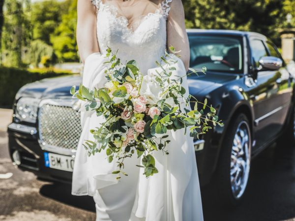 Bride in white gown holding a bouquet near a luxury car, symbolizing elegance and romance.